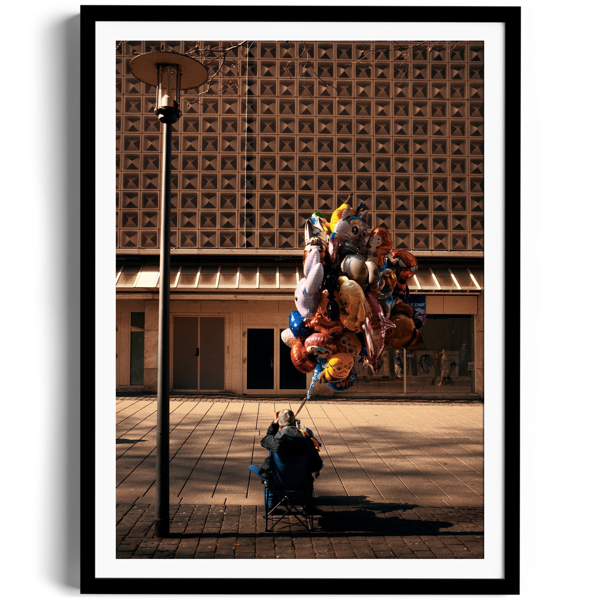 A person seated under a lamp post with a colorful assortment of balloons in a city plaza setting.