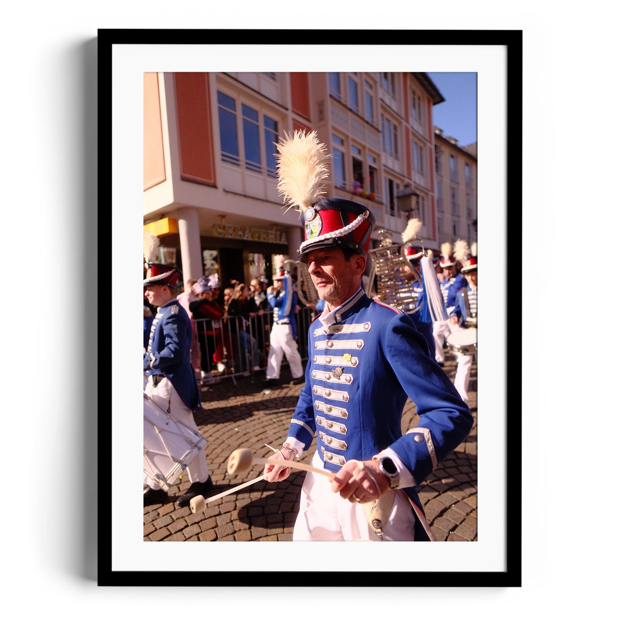 A drummer in a blue uniform with a plume hat marching in a parade, framed artwork capturing festive celebration.