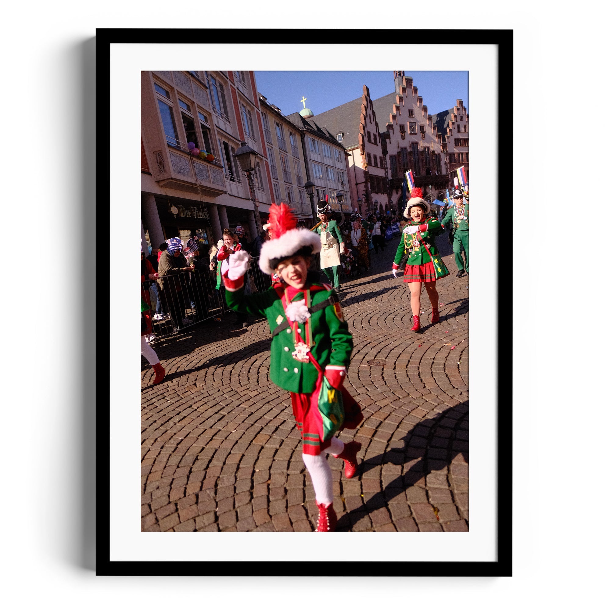 Child in green festive outfit waving at parade in a historic town square, framed fine art print.