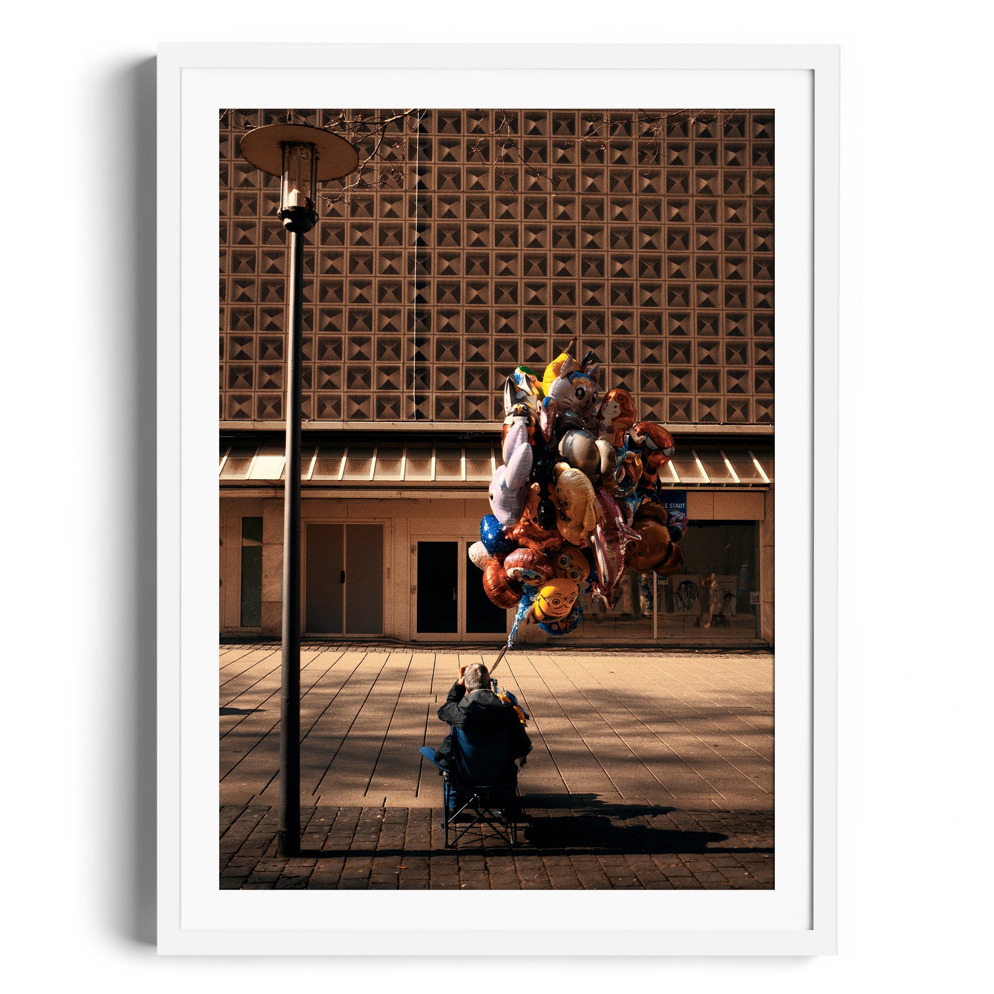 A framed fine art photograph featuring a person sitting with colorful balloons in a city plaza.